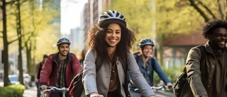 Eine Gruppe von Erwachsenen fährt gemeinsam mit Fahrrädern durch eine baumgesäumte Strasse in der Stadt. Im Vordergrund lächelt eine Frau mit lockigem Haar und Fahrradhelm in die Kamera. Die übrigen Radfahrer folgen ihr und wirken ebenfalls fröhlich.