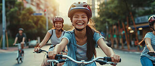 Zwei junge Frauen mit Fahrradhelmen fahren lachend auf Fahrrädern durch eine sonnige Stadtstrasse. Im Hintergrund sind weitere Radfahrer, Gebäude und Bäume zu sehen.