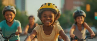 Ein fröhliches Mädchen mit gelbem Fahrradhelm fährt auf einem Fahrrad und blickt direkt in die Kamera. Hinter ihr fahren drei weitere Kinder, ebenfalls mit Helmen.